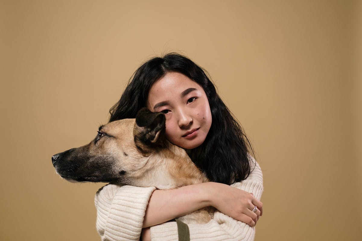 A woman with long dark hair hugs a tan dog while looking at the camera. The background is plain beige, and she is wearing a cream-colored sweater.